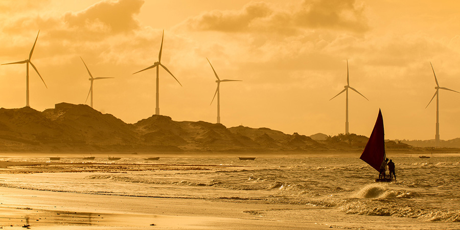 Paisagem costeira ao entardecer com turbinas eólicas em colinas ao fundo e um barco tradicional navegando nas ondas, destacando a integração de energia renovável com a natureza.