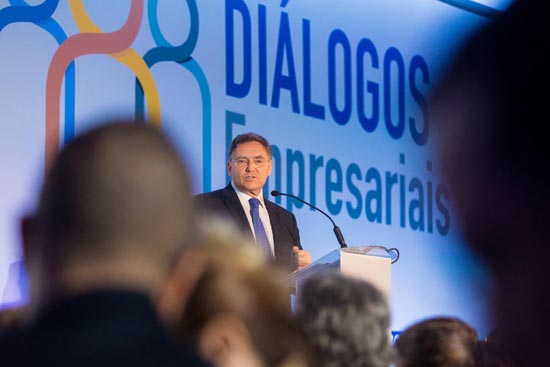 Indivíduo no pódio durante o evento 'DIÁLOGOS Empresariais,' com público corporativo ao fundo, destacando discussões sobre negócios e liderança.