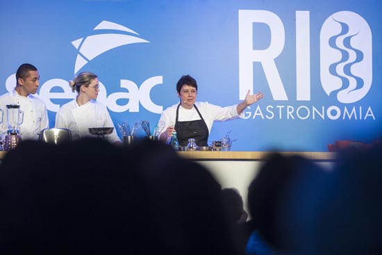 Chefs apresentando uma demonstração culinária no evento Rio Gastronomia, com ingredientes e equipamentos exibidos em um balcão.