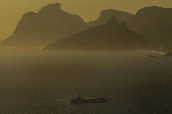 Imagem de um navio navegando em um amplo corpo d'água ao pôr ou nascer do sol, com montanhas escuras ao fundo e luz dourada criando um efeito atmosférico.