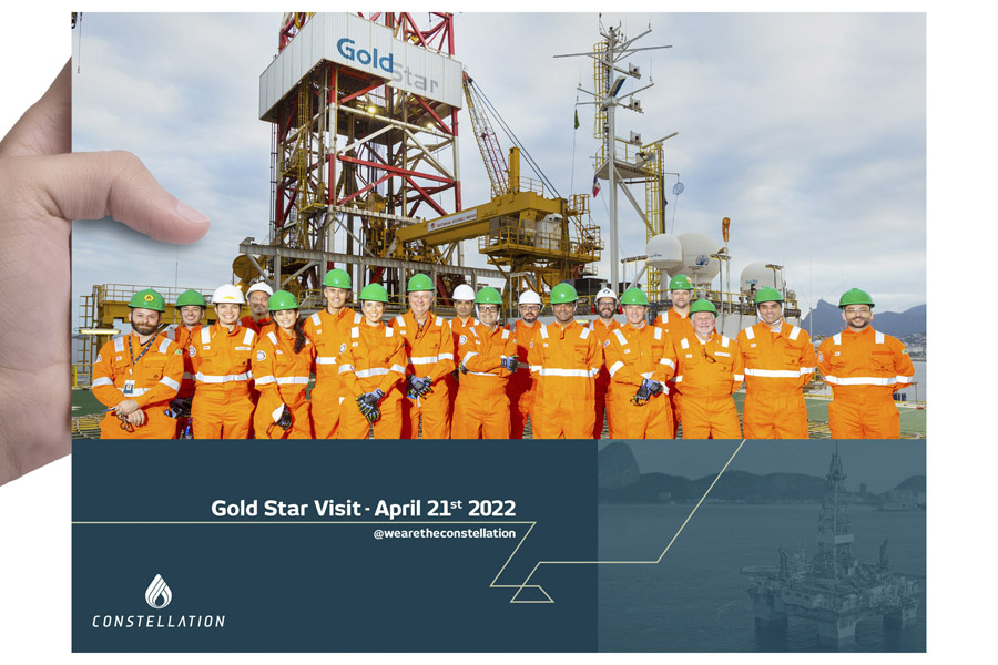 Grupo em uniformes de segurança laranja e capacetes posando para uma foto em frente à instalação industrial chamada 'Gold Star', com destaque para o logotipo 'CONSTELLATION'.